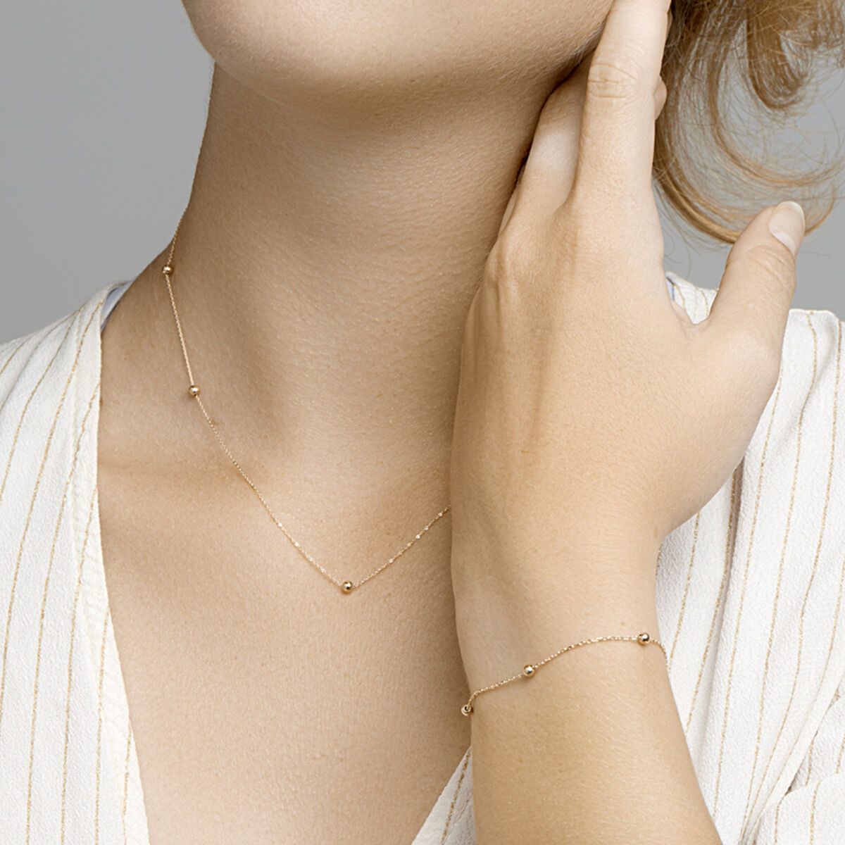 Een vrouw modelleert de Van der Kooij Collectie armband anker en bolletjes 1,0 mm 17,5-18,5cm in 14K geelgoud, gecombineerd met een bijpassende ketting en een lichte V-hals blouse, die haar hals zachtjes raakt.