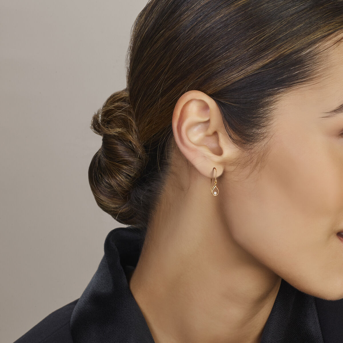 Een vrouw met donker haar in een lage knot draagt een Van der Kooij Collectie oorhangers met Franse haak en zirkonia 23 mm van 14K geelgoud. Ze is gekleed in een zwarte top en de afbeelding benadrukt haar oor en profiel tegen een lichtbeige achtergrond.