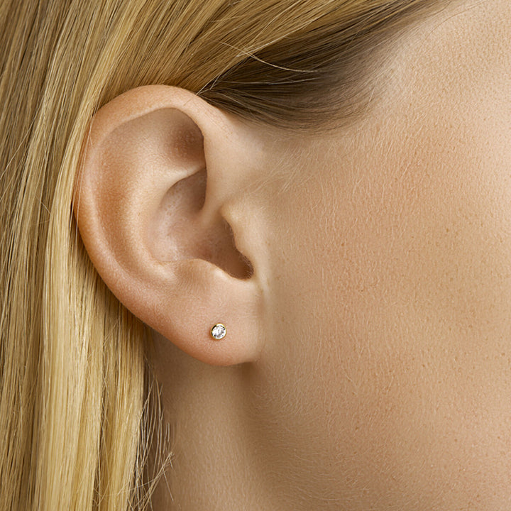 Close-up van het oor van een persoon met steil blond haar, gekleed in Van der Kooij Collectie oorknoppen zirkonia 14K geelgoud - kleine ronde oorbellen bezet met een heldere zirkonia edelsteen.