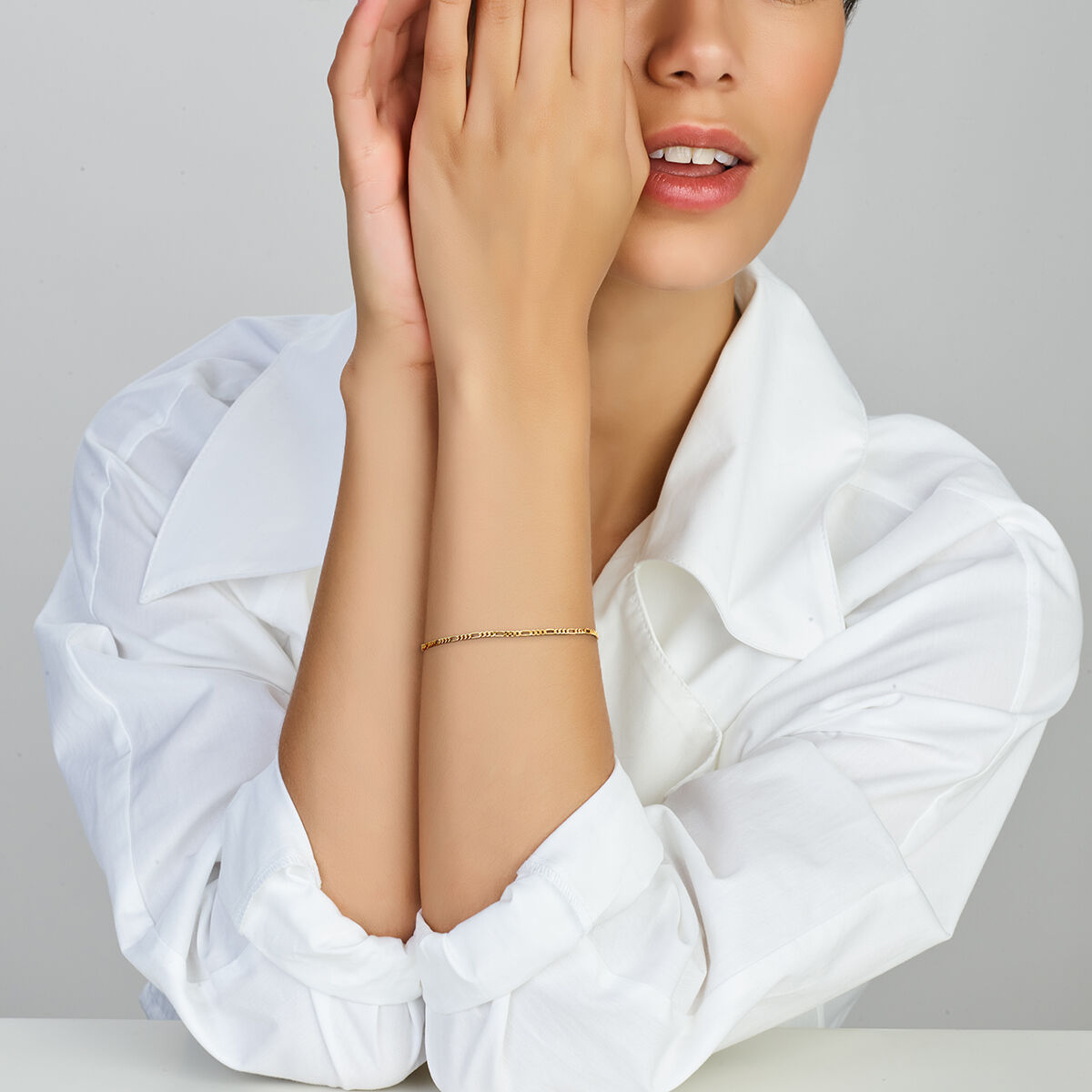 Een vrouw in een wit overhemd poseert met één hand gedeeltelijk voor haar gezicht, en toont de Gouden Armband Dames Figaro 14K uit de Van der Kooij Collectie. Ze heeft kort haar, neutrale make-up en zit tegen een effen achtergrond.