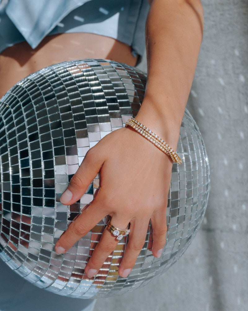 Een close-up van een hand versierd met de Excellent Jewelry Geelgouden Tennisarmband met 3.00 Karaat Diamanten in Chatonzetting - 14 Karaat Goud, rustend op een reflecterende discobal, gepaard met ringen tegen een heldere achtergrond en lichtblauwe kleding.