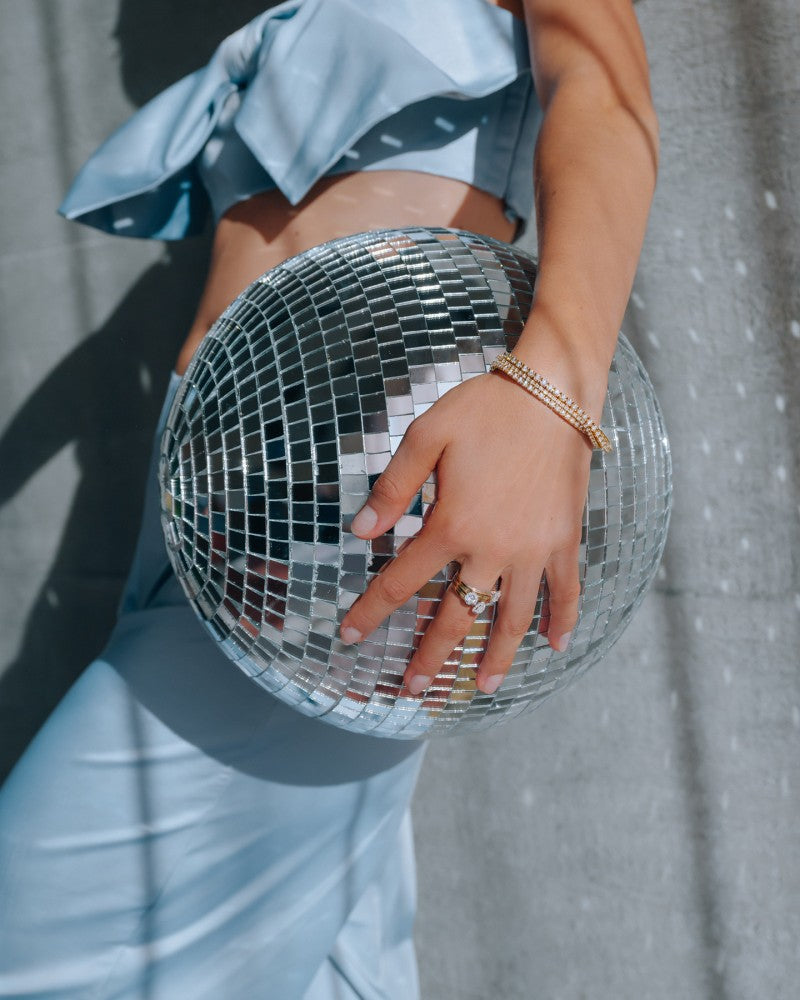 Een persoon in een lichtblauwe outfit en sieraden, waaronder de Excellent Jewelry Geelgouden Tennisarmband met 3.00 karaat diamanten in chatonzetting, houdt een reflecterende discobal vast terwijl zonlicht schaduwen werpt op een muur met grijze textuur.