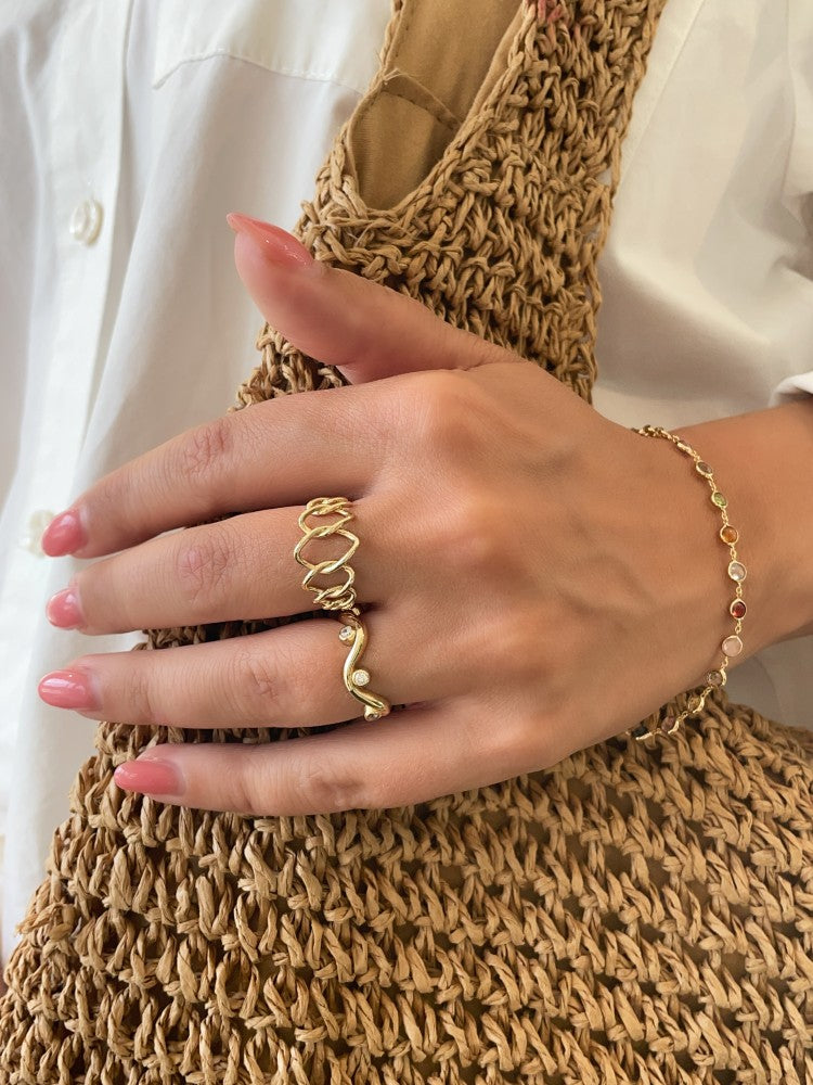 Een gemanicuurde hand draagt twee gouden ringen, een geelgoud edelsteen armband met veelkleurige stenen en houdt een geweven rieten tas vast. De persoon is gekleed in een wit overhemd met knoopsluiting.