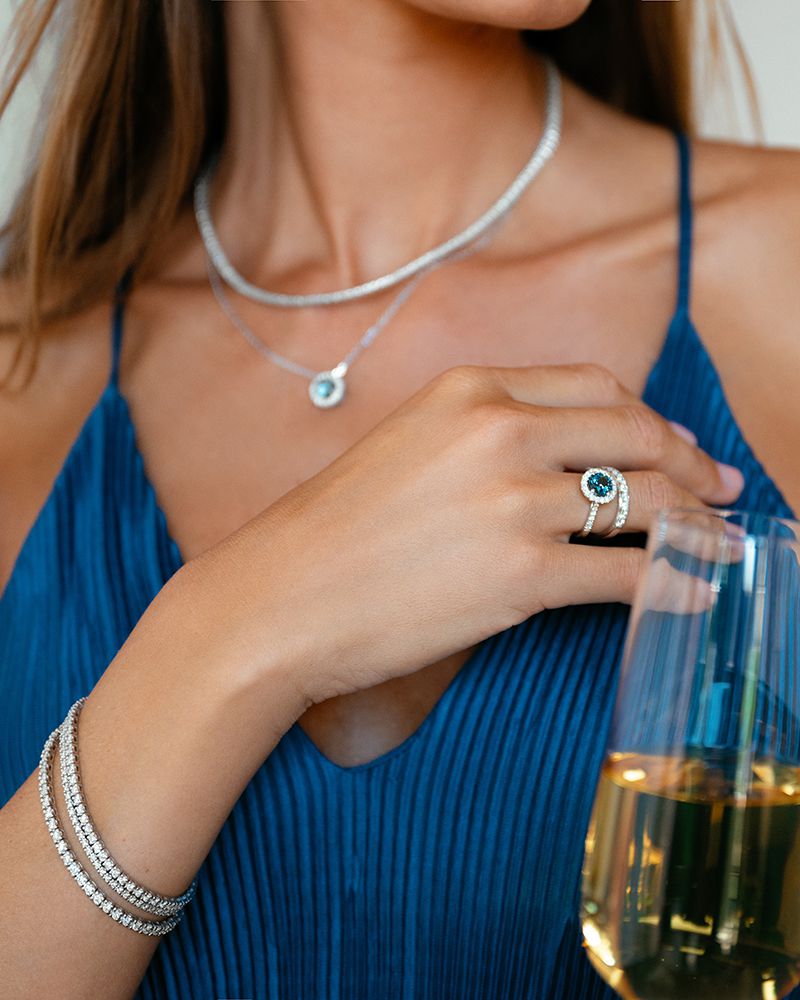 Een vrouw in een blauwe geplooide jurk houdt een glas witte wijn vast. Ze draagt een zilveren hanger ketting, een smaragd en diamanten ring, en twee Excellent Jewelry Witgouden Armbanden met Diamanten - 2.01 crt Briljant, 14 Karaat.