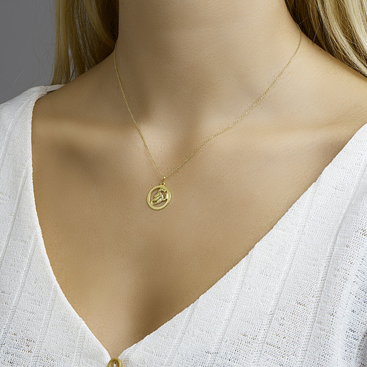 Een vrouw draagt een witte, gestructureerde V-hals top en een gouden collier met het Van der Kooij Collectie hanger sterrenbeeld gediamanteerd 14K geelgoud, met een verhoogd motief, afgebeeld van schouders tot borst.