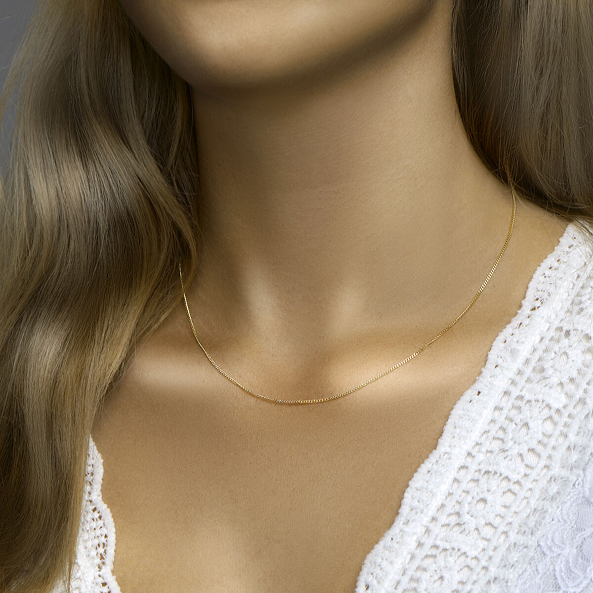 Een vrouw met lang lichtbruin haar draagt een Van der Kooij Collectie collier gourmette 4-zijdes geslepen 1,2 mm 41 + 4 cm 3 micron zilver verguld (geel) en een wit kanten topje, afgebeeld vanaf de schouders omhoog.