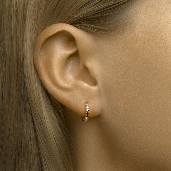 Close-up van het oor van een vrouw met Van der Kooij Collectie klapoorbellen 14K geelgoud met zwarte en gouden kraaltjes. Haar steile blonde haar is weggestopt achter haar oor, tegen een neutrale achtergrond.