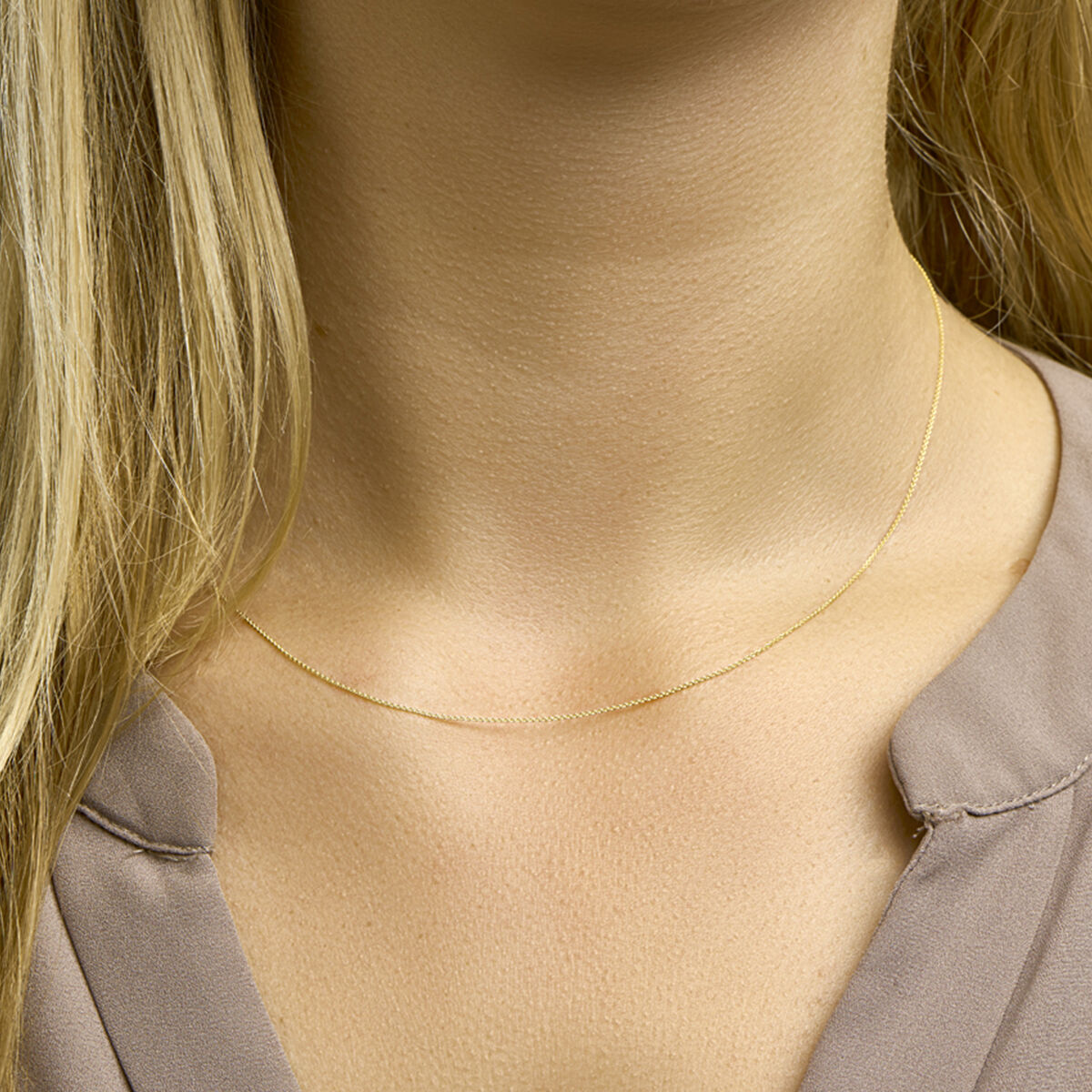 Een close-up van de hals van een vrouw met een delicaat collier anker plat 1,0 mm 14K geelgoud van Van der Kooij Collectie, met blond haar en een taupe blouse waarvan de halslijn met inkeping gedeeltelijk zichtbaar is.
