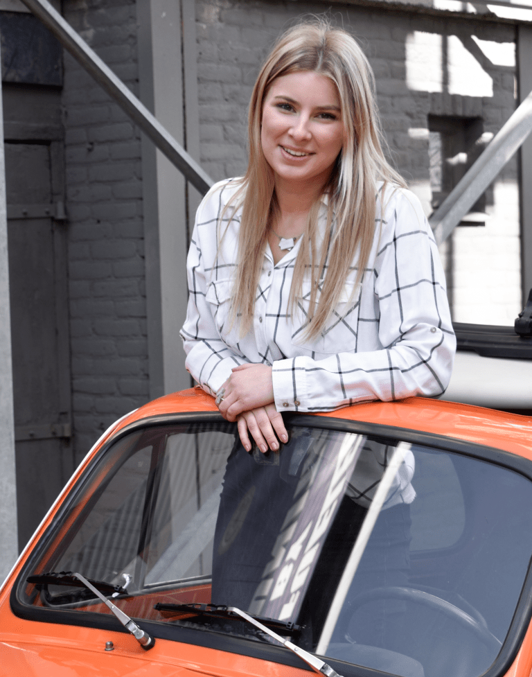 Een persoon met lang blond haar, gekleed in een wit geblokt shirt, lacht terwijl hij tegen de voorruit van een oranje auto leunt. Ze staan voor een bakstenen gebouw met metalen structuren, gekleed in CLIC Creations Collier gekruiste rechthoeken & hematiet C233.