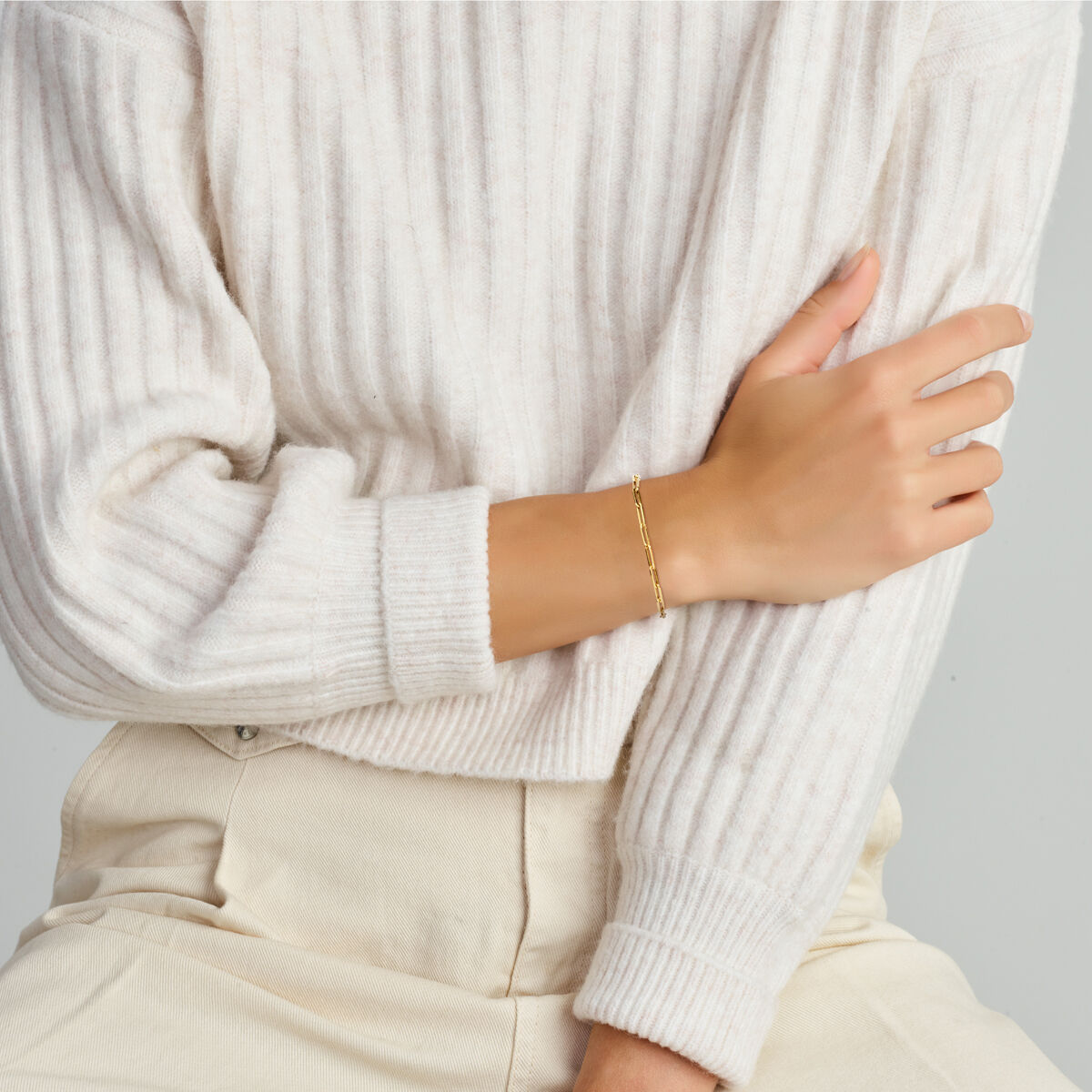 Een persoon in een crèmekleurige geribbelde trui en lichte broek zit met één arm gekruist en toont de Van der Kooij Collectie armband paperclip vierkante buis 3,0 mm 18 cm met groot springslot in 14K geelgoud.