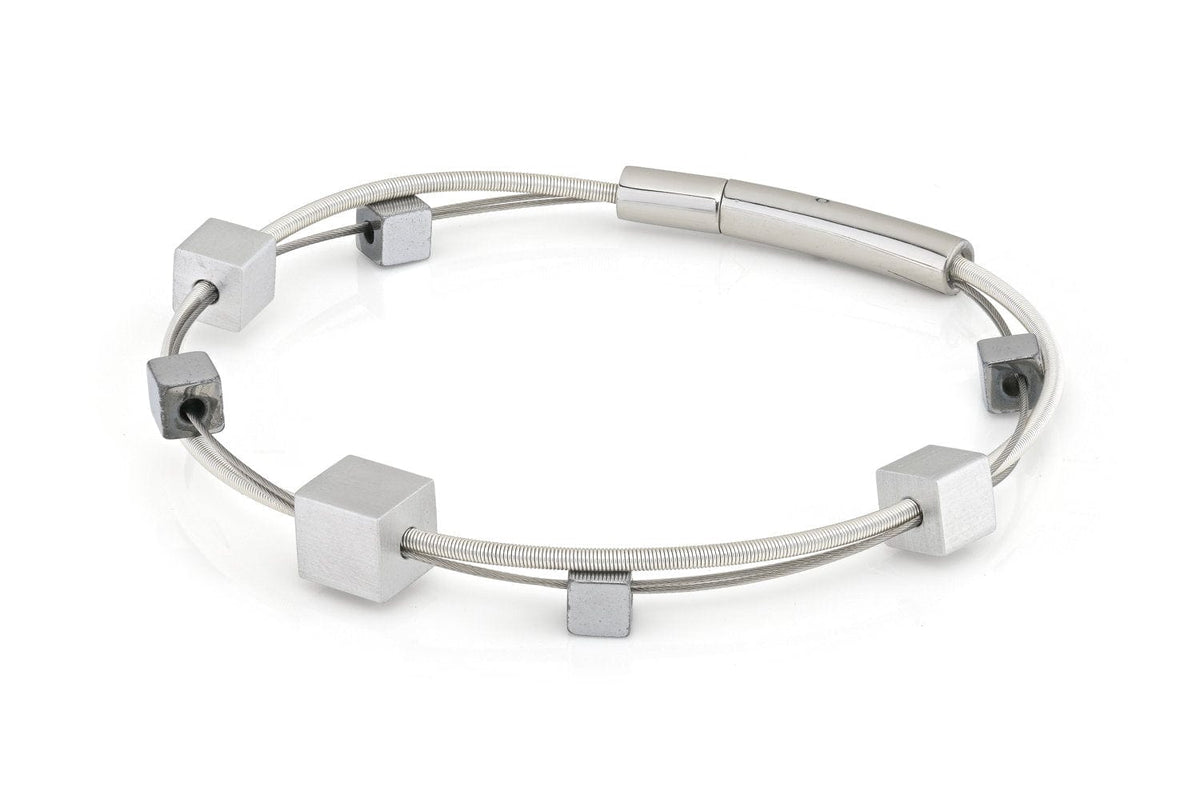 De CLIC Creations Armband speelse kubusjes zilver A257 is een minimalistische zilveren armband, gemaakt van hypoallergene materialen, met een strak, dun draadontwerp versierd met gelijkmatig verdeelde vierkante metalen kralen en een cilindervormige sluiting.
