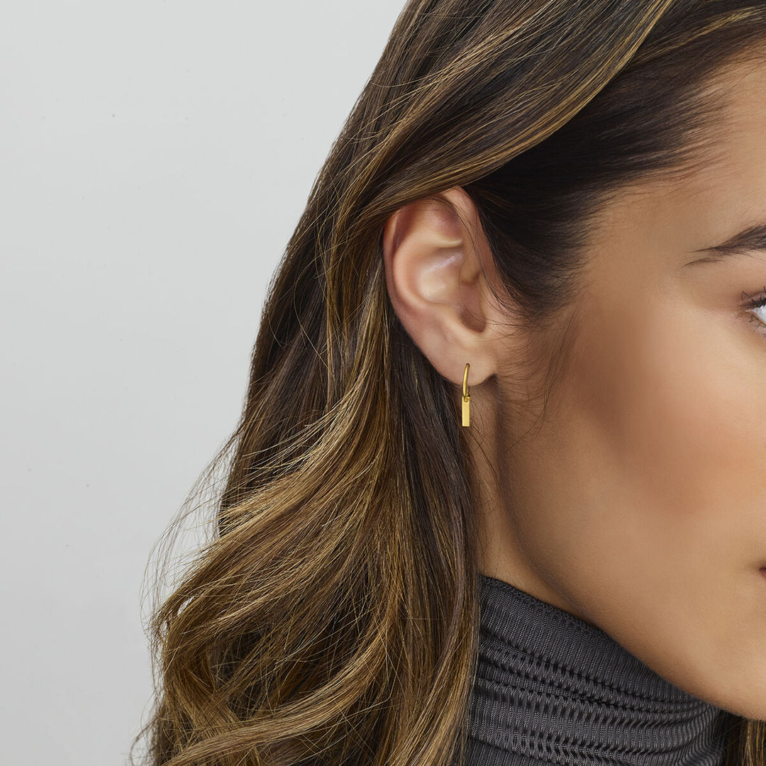 Een close-up van het oor van een vrouw toont Van der Kooij Collectie oorbellen plaatje 14K geelgoud-kleine rechthoekige geelgouden hoepels. Ze heeft lang gehighlight bruin haar en draagt een geribbelde grijze coltrui op een lichtgrijze ondergrond.