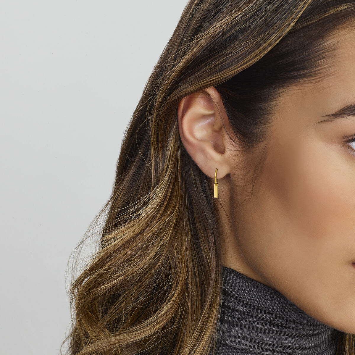 Een close-up van het oor van een vrouw toont Van der Kooij Collectie oorbellen plaatje 14K geelgoud-kleine rechthoekige geelgouden hoepels. Ze heeft lang gehighlight bruin haar en draagt een geribbelde grijze coltrui op een lichtgrijze ondergrond.