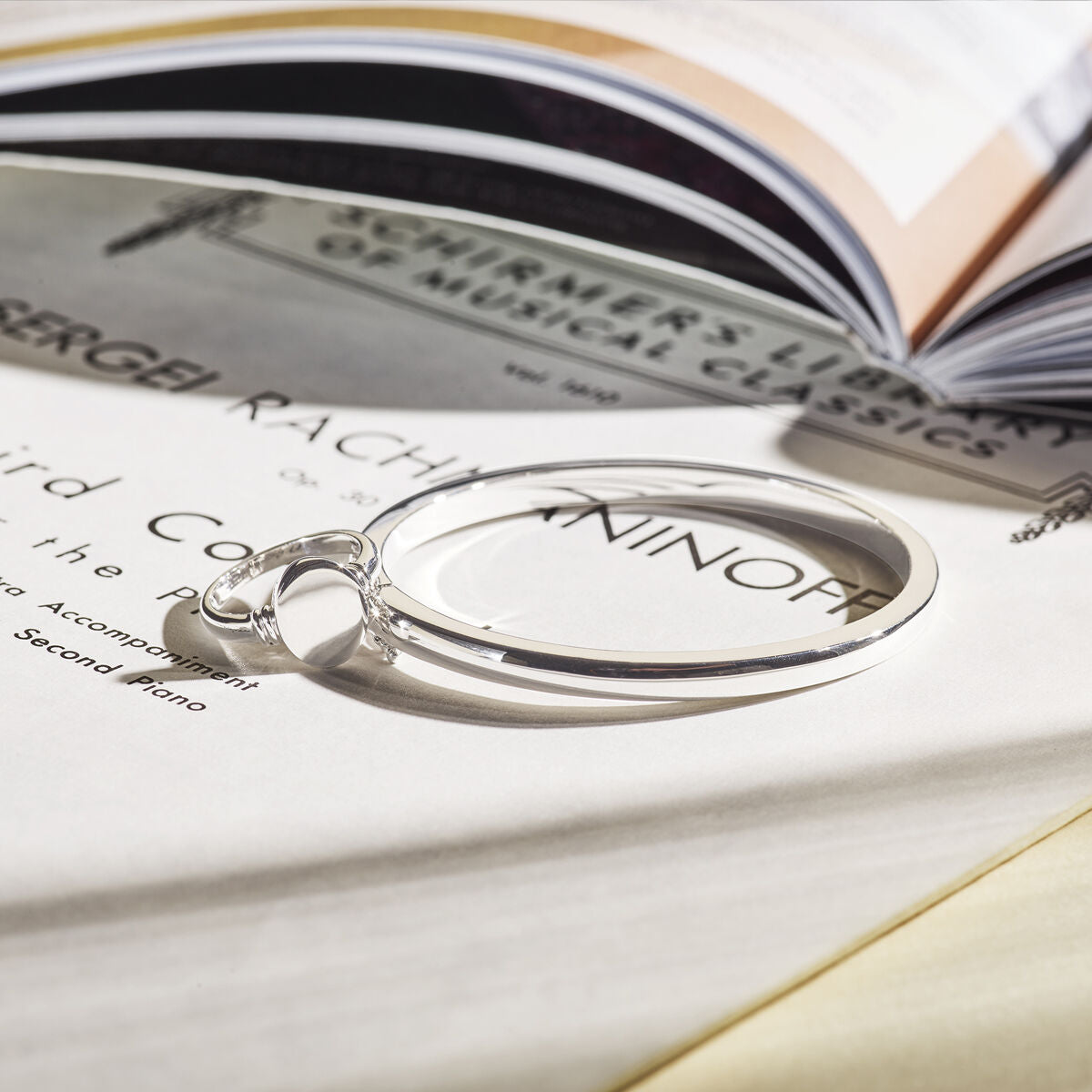 De bangle scharnier 4 mm bolle buis zilver gerhodineerd van Van der Kooij Collectie ligt op open bladmuziek en een boek, terwijl zacht licht subtiele schaduwen werpt.