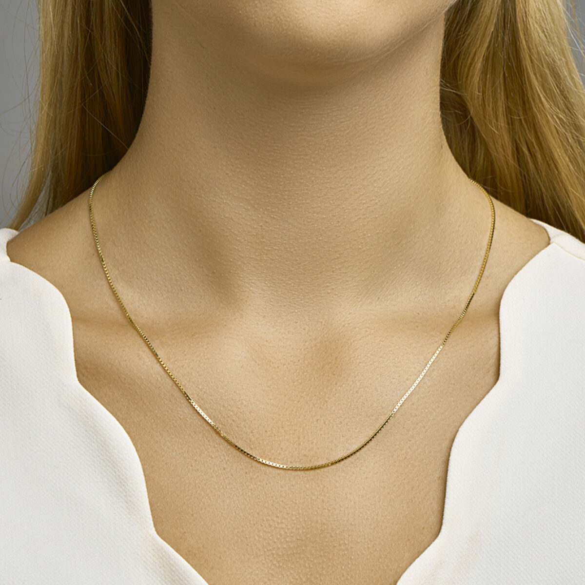 Een vrouw draagt een Van der Kooij Collectie collier venetiaans 1,1 mm 14K geelgoud met een wit topje met geschulpte halslijn; de focus ligt op haar nek en de delicate gouden ketting, waardoor haar gezicht uit het zicht blijft.