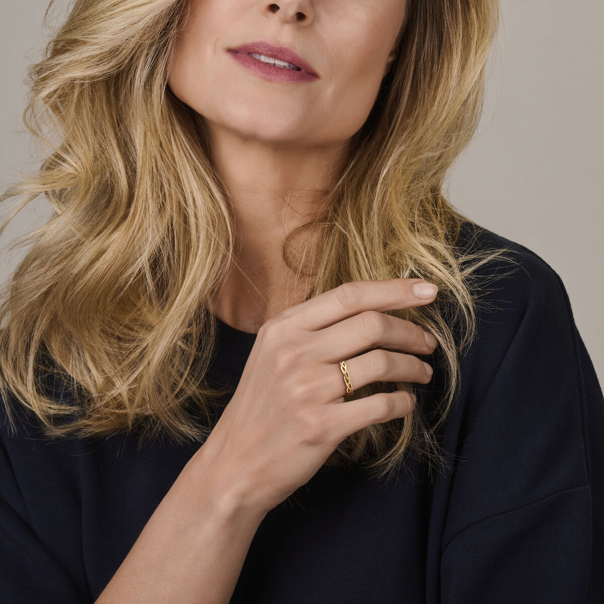 Een vrouw met golvend blond haar in een donker topje poseert met haar hand voor haar gezicht. Ze showt de Van der Kooij Collectie ring gevlochten 14K geelgoud - tijdloze elegantie in een gevlochten gouden ontwerp.