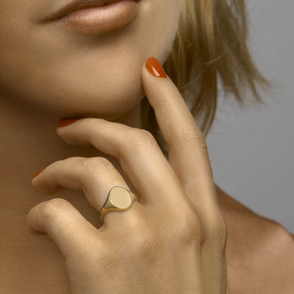 Een close-up van een vrouw met een lichte huidskleur die de Van der Kooij Collectie graveerring ovaal 14K geelgoud aan haar wijsvinger draagt en haar gezicht aanraakt. Ze heeft kort, lichtbruin haar en oranje nagellak.