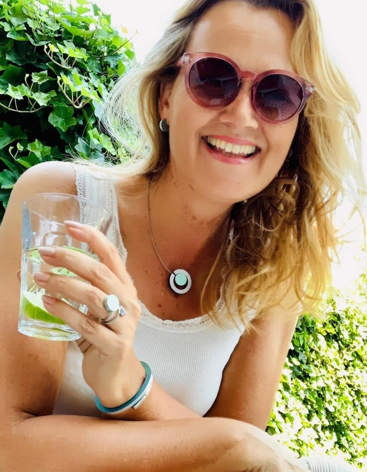 Een vrouw met lang blond haar en een zonnebril lacht terwijl ze een glas met een schijfje limoen vasthoudt. Ze draagt een wit tanktopje, oorringen en een ronde ketting en draagt de Ring Twee ronde vormen Goud|Zand R5G van CLIC Creations. Groen blad accentueert het handgemaakte tafereel uit Nederland.