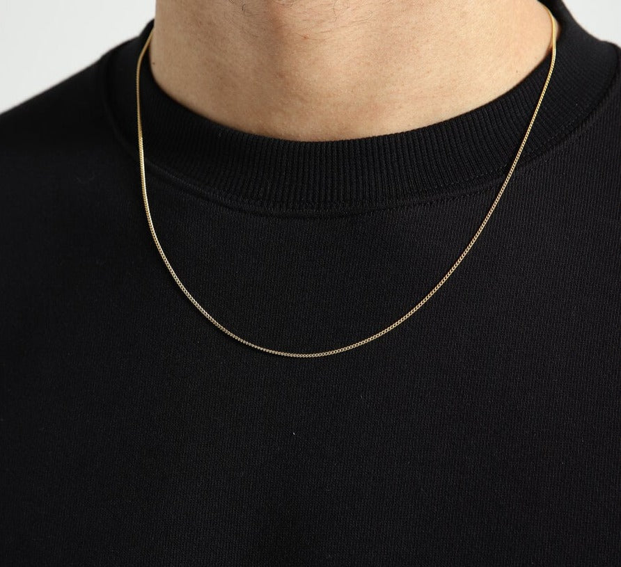 Een persoon die de Juwelier van der Kooij Goudkleurige heren ketting gourmette - 1,4 mm zilver verguld draagt over een zwart shirt met ronde hals, met focus op de ketting en het bovenste deel van de borst.