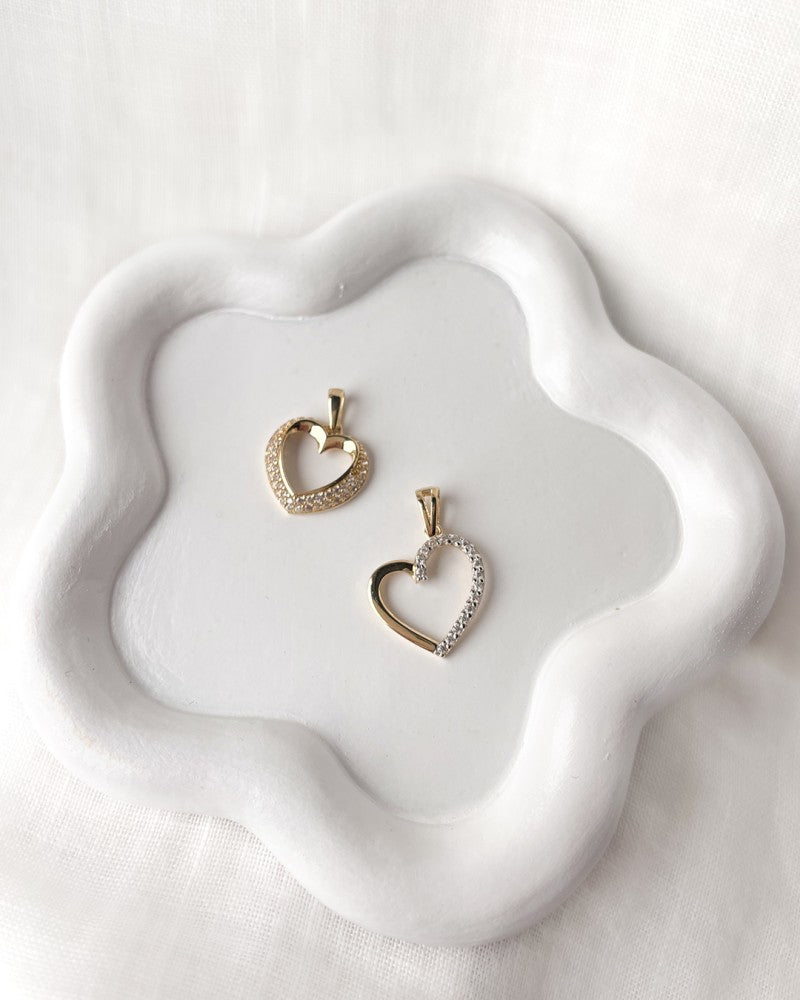 Een 14 karaat gouden harthanger met bol design, gezet met zirkonia's (1,0 gram, 14 mm) van Excellent Jewelry roest op een witte keramische schaal met golvende rand op een zachte witte stoffen achtergrond.