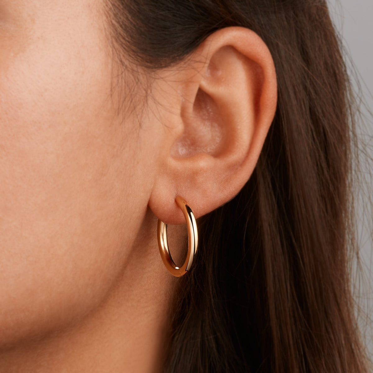 Close-up van het oor van een vrouw met Boccia Titanium Oorringen gp 05072-02 van Boccia, met glanzende gouden oorringen. Haar steile bruine haar is gedeeltelijk zichtbaar tegen een neutrale achtergrond.