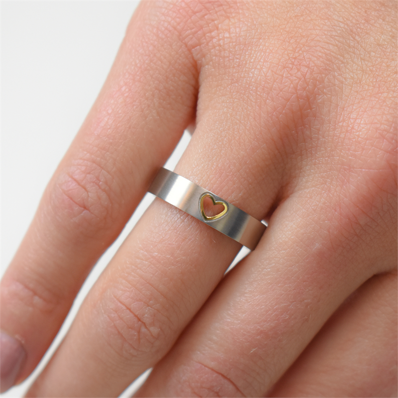 Een close-up van een hand die de Boccia Titanium Ring Bicolor 0143-02 van Boccia draagt, met een klein gouden hartje in het midden op de middelvinger tegen een effen witte achtergrond.