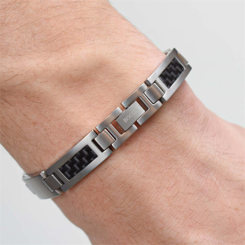 Een close-up afbeelding toont een lichtgekleurde pols met een Boccia Titanium Armband carbon 0333-01 van Boccia. Gemaakt van strak titanium, heeft het zwarte rechthoekige carbon inzetstukken en gegraveerde tekst, wat het moderne, gesegmenteerde ontwerp benadrukt tegen een effen achtergrond.