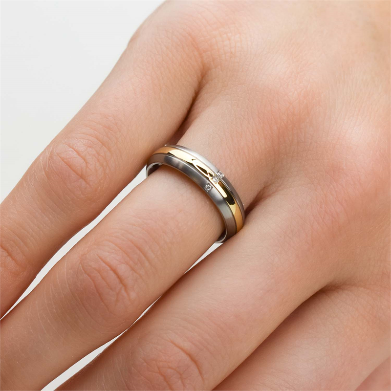 Een close-up toont een hand die de Boccia Titanium Ring Bicolor dia 3/0.015 - 0131-04 van Boccia draagt, in goud en zilver met een kleine centrale diamant, aan de ringvinger tegen een effen lichte achtergrond.