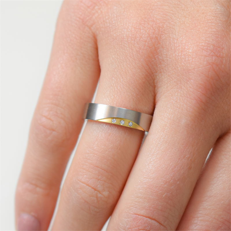 Een close-up van een hand die de Boccia Titanium Ring Bicolor dia 3/0.015 - 0138-04 van Boccia draagt, met drie kleine diamanten op een rij op de mat zilveren en gouden band, gedragen aan de middelvinger.