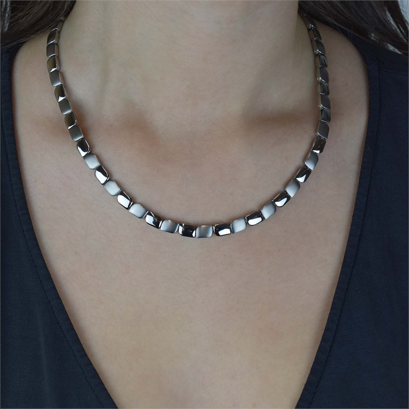 Een persoon draagt de Boccia Titanium Collier 08003-01, een glanzende, zilverkleurige halsketting met vierkante schakels van Boccia. Het ligt plat tegen de huid, buigt soepel rond de nek en past goed bij een donker topje met V-hals.