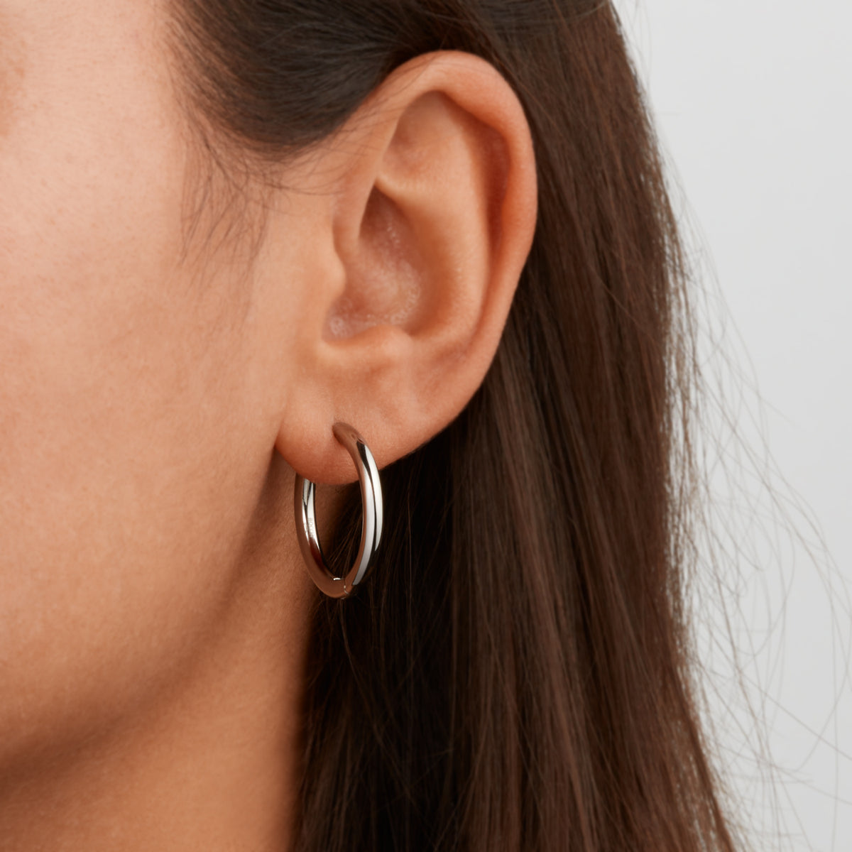 Close-up van een oor met een eenvoudige zilveren Boccia Titanium Oorringen 05072-01 creolen met een klapsluiting, die contrasteert met het donkere, steile haar van de persoon. De effen, lichtgekleurde achtergrond versterkt de elegantie van deze Boccia titanium oorbellen.
