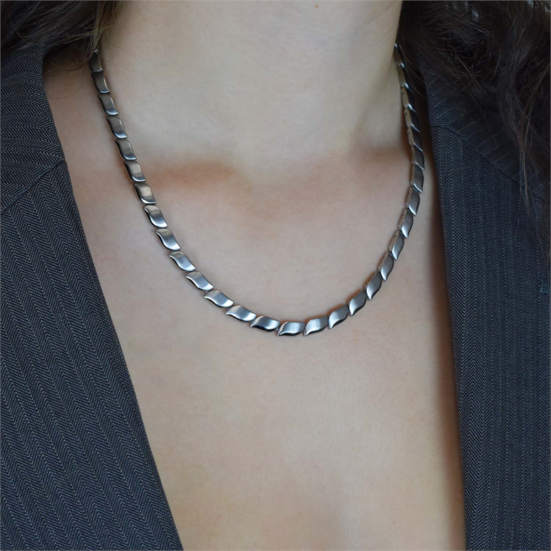 Een persoon draagt de elegante Boccia Titanium Collier 08038-01, een zilverkleurige damesketting met een visgraatmotief, gecombineerd met een donkere, getextureerde blazer. De platliggende ketting contrasteert prachtig tegen hun huid in een neutrale, soft-focus achtergrond.