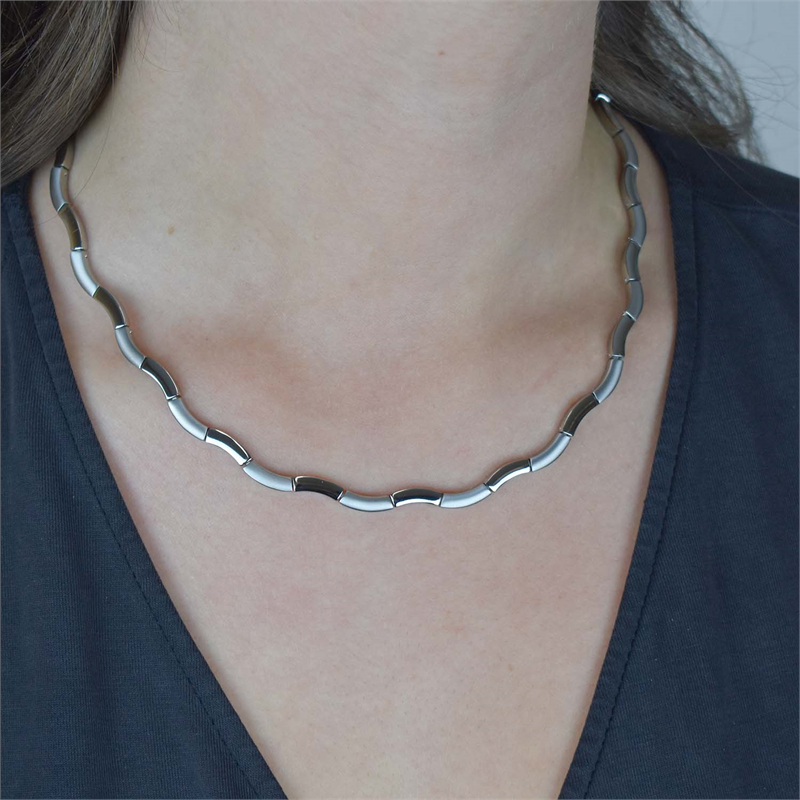 Een close-up van een persoon die de Boccia Titanium Collier 0844-01 draagt, een zilveren, gesegmenteerde ketting met een golvend ontwerp, afgebeeld op de huid en een donker topje met V-hals.