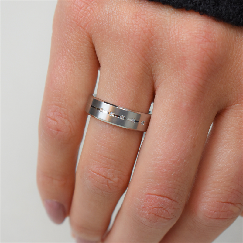Een close-up van een hand die de Boccia Titanium Ring dia 5/0.025 - 0101-20 draagt, met kleine vierkante uitsparingen en een geborstelde afwerking op de ringvinger, met de persoon in een zwarte hoes.