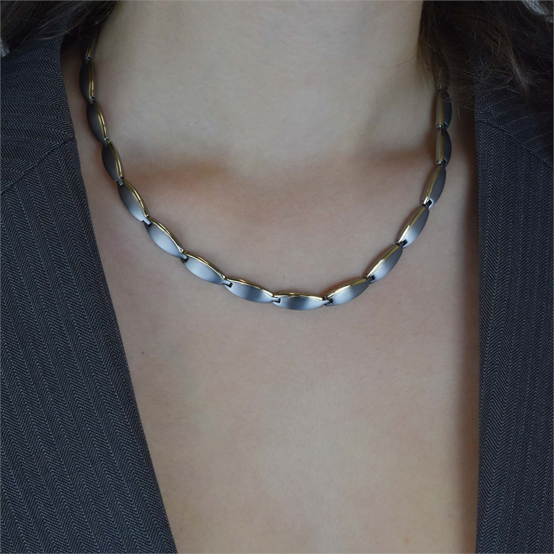 Een close-up van een persoon die de Boccia Titanium Collier Bicolor 08034-02 draagt, met ovaalvormige geborstelde bicolor schakels, afgebeeld tegen een blote huid en een donkere krijtstreepblazer voor een elegante look.