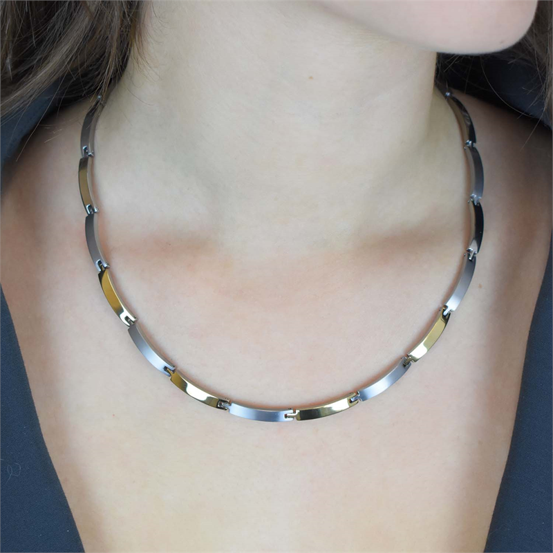 Een close-up van een persoon die de Boccia Titanium Collier Bicolor 0816-03 draagt, met afwisselend zilveren en gouden rechthoekige titanium schakels, tegen een achtergrond van blote huid en een donker, openvallend bovenstuk.