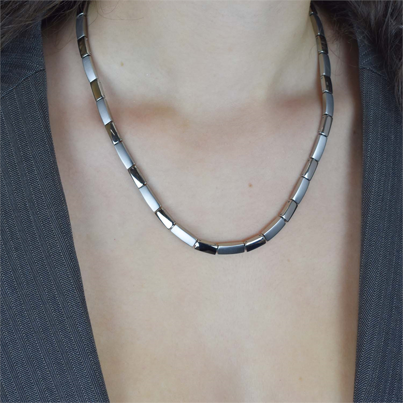 Een persoon in een grijs gebloemd jasje draagt de elegante Boccia Titanium Collier NL 08043-01, met een strak, metallic ontwerp met rechthoekige schakels. De afbeelding benadrukt de ketting en het bovenste deel van de borst.