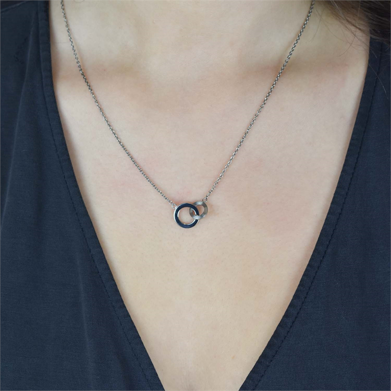 Een close-up van een persoon die de Boccia Titanium Figura Collier 08033-01 van Boccia draagt, met een hanger van in elkaar grijpende cirkels, afgebeeld tegen hun blote nek en een donker topje met V-hals.