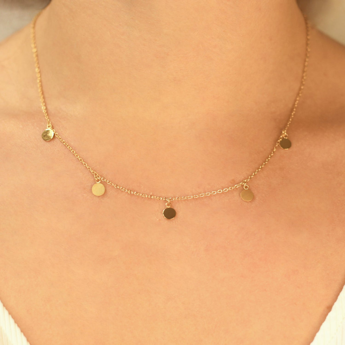 Een close-up van een persoon die de Boccia Titanium Collier gp 08064-02 van Boccia draagt, een delicate goudkleurige ketting met kleine, ronde, platte bedeltjes gelijkmatig verdeeld langs de ketting, die net boven het sleutelbeen rust.