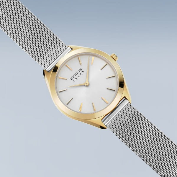 De Bering 17331-010 dameshorloge heeft een gouden en zilveren ontwerp, witte wijzerplaat, gouden uurmarkeringen, zilveren mesh band en saffierglas, afgebeeld tegen een lichtblauwe achtergrond met kleurverloop.