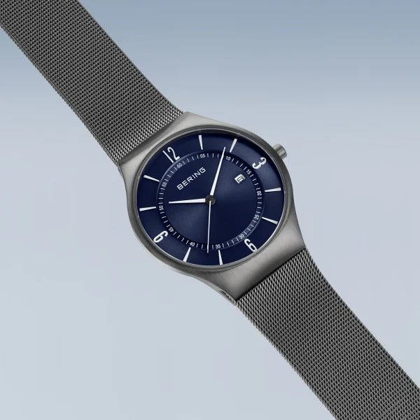 De Bering 15940-378 heeft een zilverkleurige mesh band, blauwe wijzerplaat, saffierglas, zilverkleurige uurmarkeringen en wijzers en een datumweergave, allemaal tegen een lichtblauwe gradiënt achtergrond.