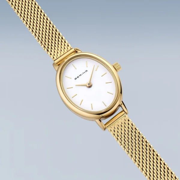De Bering 11020-334 is een goudkleurige dameshorloge met mesh band, ovaal witte wijzerplaat en minimalistisch design. Het quartz uurwerk heeft gouden uurmarkeringen op een gradiënt lichtblauwe achtergrond.