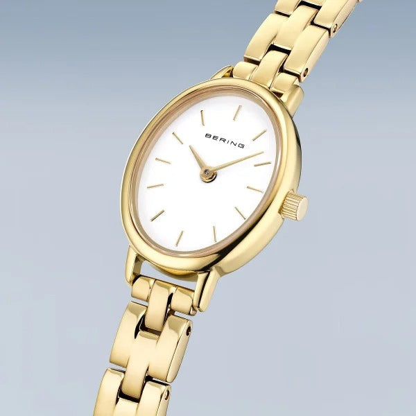 Het Bering dameshorloge 11020-734 heeft een goudkleurige schakelband, witte wijzerplaat en minimalistische uurmarkeringen. Dit stijlvolle horloge beschikt over een betrouwbaar quartz uurwerk en is afgebeeld tegen een lichtblauwe achtergrond.