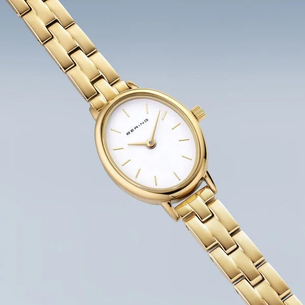 De dameshorloge Bering 11020-734 in goudkleur met wit ovaal wijzerplaat, gouden urenmarkeringen en schakelband wordt getoond op een lichtblauwe achtergrond. Het quartz uurwerk en de merknaam Bering completeren het ontwerp.