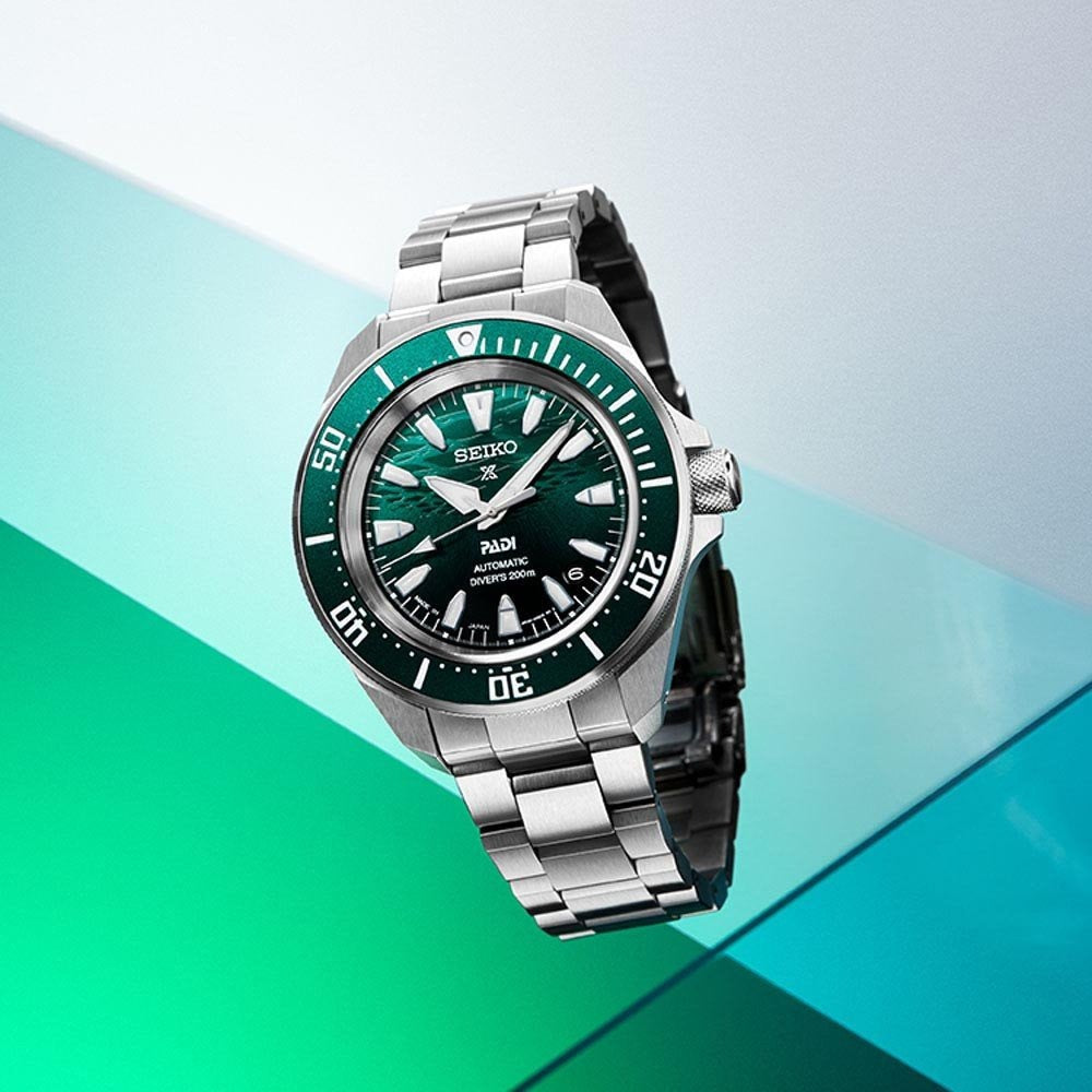 De Seiko heren horloge SRPL53K1 heeft een groene wijzerplaat en lunette, een zilverkleurige roestvrijstalen band en opvallende uurmarkeringen op een geometrische groenblauwe en witte achtergrond.