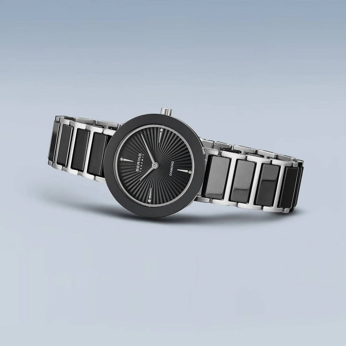 Het Bering dames horloge 11429-702 heeft een zwarte en zilveren band, ronde zwarte wijzerplaat met minimalistische indexen, sunburst-effect en saffierglas, fraai gepresenteerd tegen een lichtblauwe achtergrond.