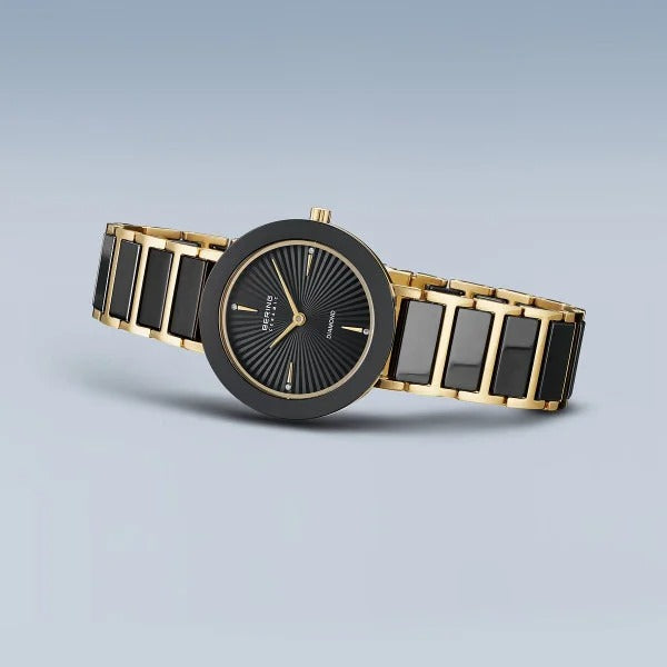 Het Bering dames horloge 11429-732 heeft een zwarte keramische en gouden schakelband, een ronde zwarte wijzerplaat met gouden wijzers en markers, plus een zonnestraalpatroon op een lichtblauwe achtergrond met kleurverloop.