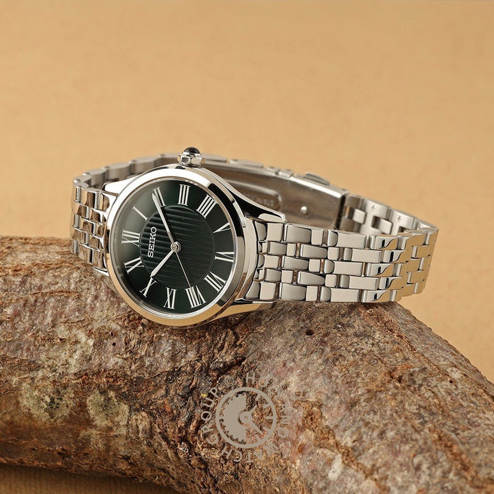 Een zilveren Seiko dameshorloge SUR611P1 met een metalen band, zwarte wijzerplaat, saffierglas en Romeinse cijfer uurmarkeringen rust op een gestructureerde bruine tak tegen een bruine achtergrond.