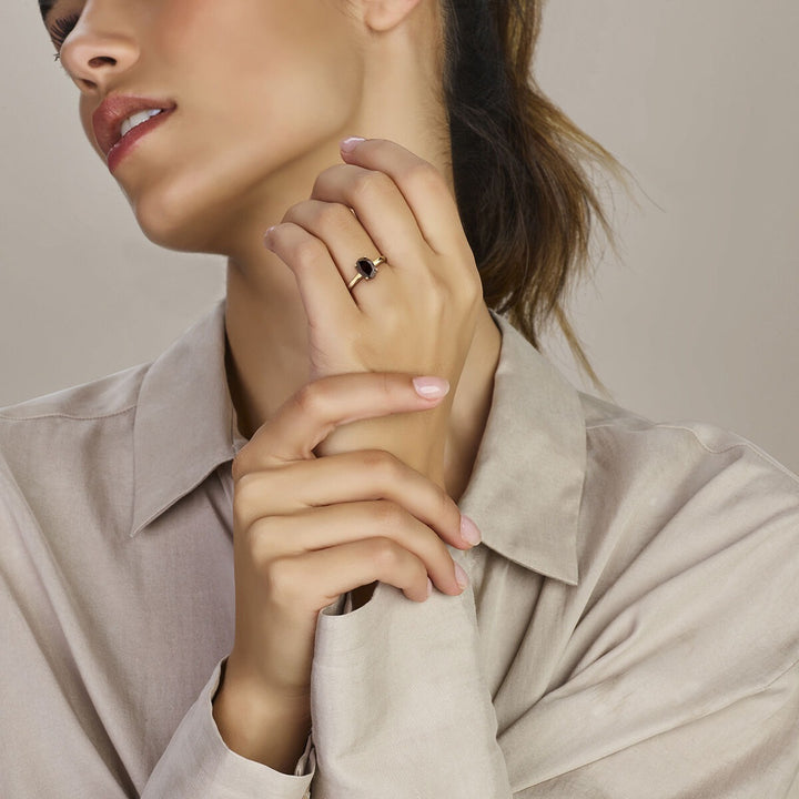 Een vrouw in een beige overhemd raakt voorzichtig haar gezicht aan en toont de Van der Kooij Collectie ring granaat 14K geelgoud. Haar nagels zijn netjes gemanicuurd in een natuurlijke tint.