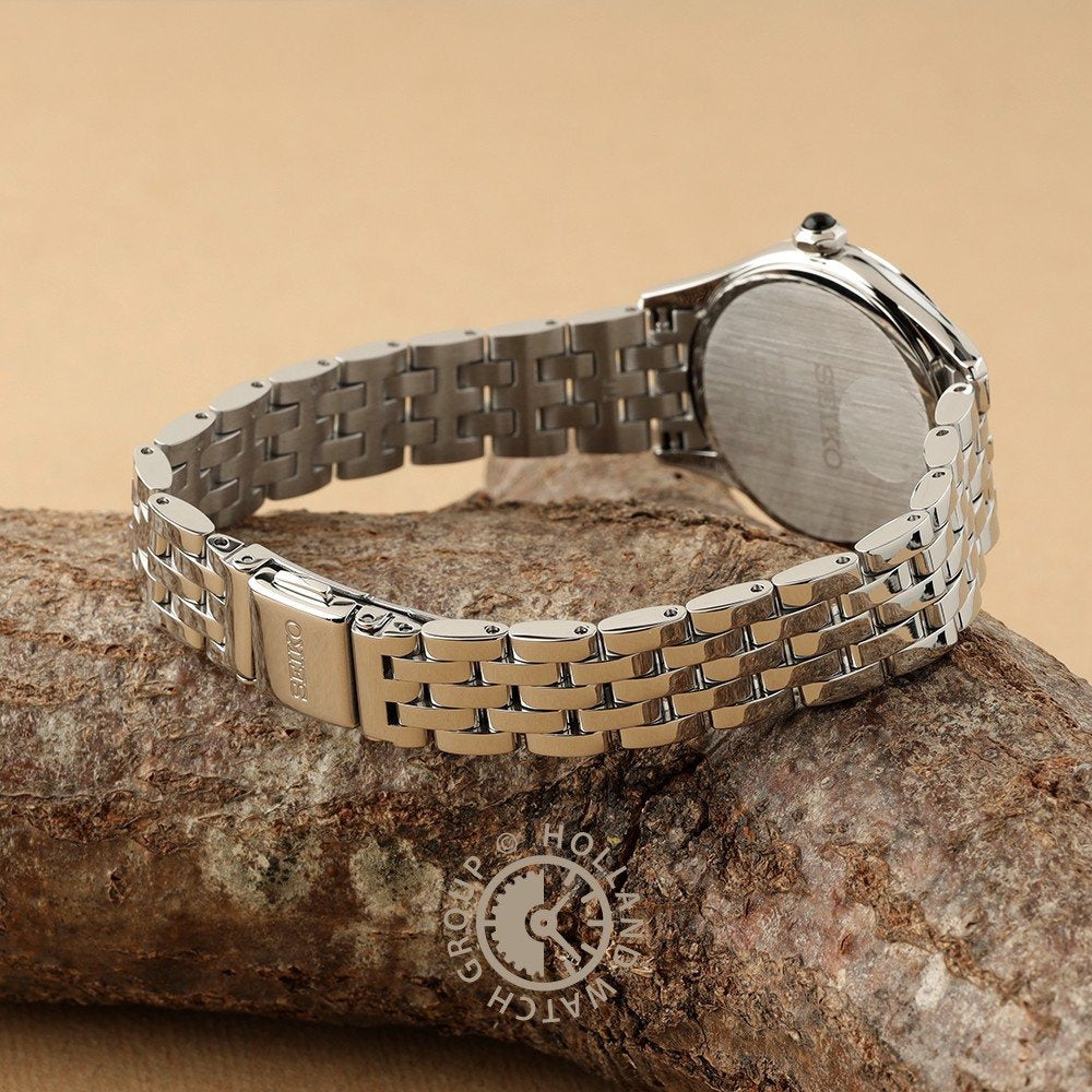 De Seiko dames horloge SUR611P1 heeft een gepolijste roestvrijstalen band en rust op bruin getextureerd hout, waardoor de elegante sluiting en achterkant van de kast zichtbaar zijn. Het Seiko logo is zichtbaar op zowel de sluiting als de achterkant van dit stijlvolle quartz horloge.